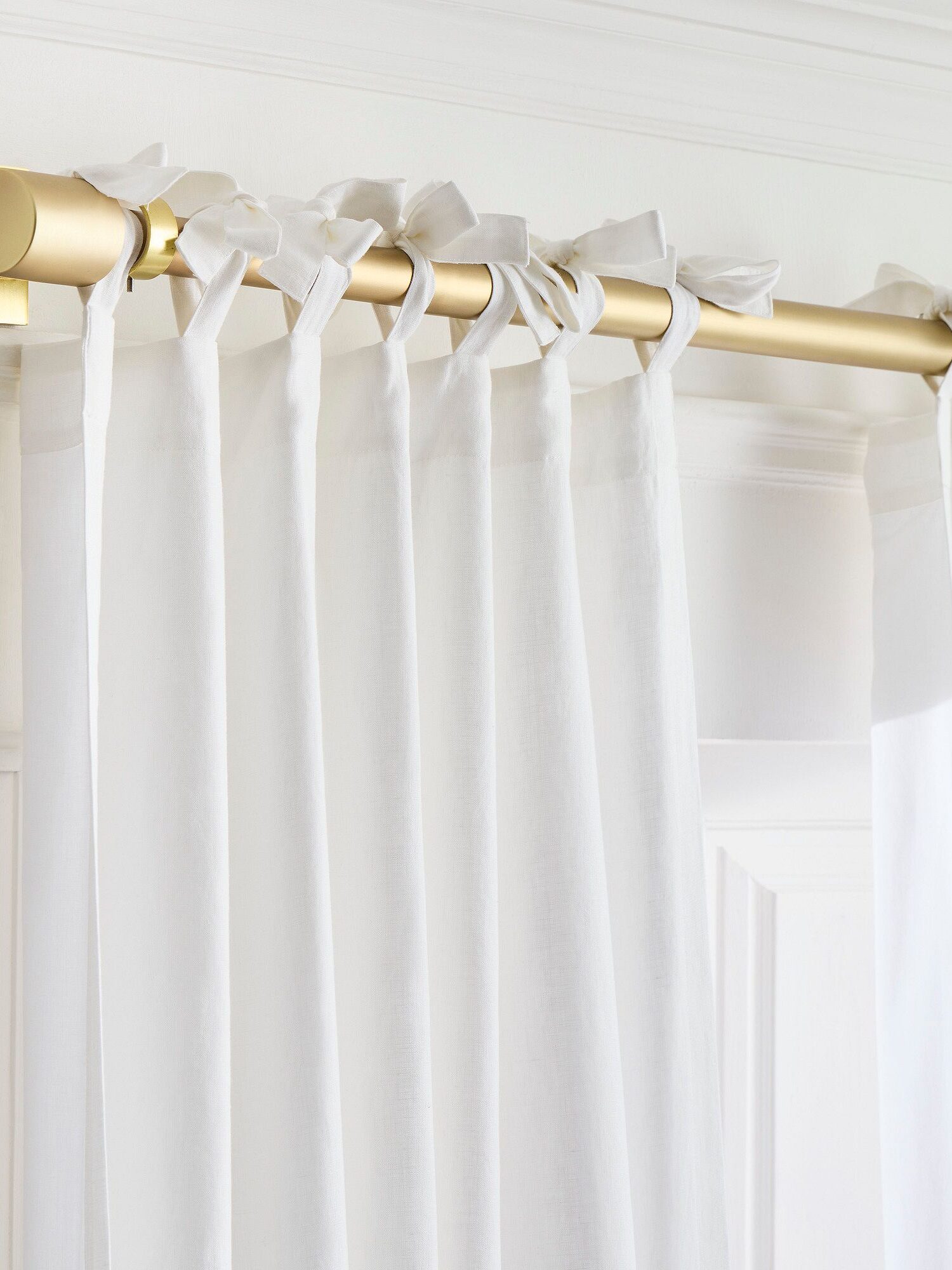 Gold curtain rod with sheer white curtains tied at the top with fabric bows, mounted on a white wall.