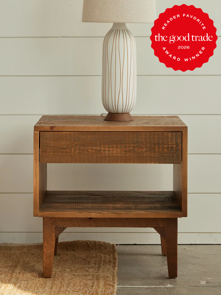 A wooden nightstand with an open shelf and a drawer, topped with a ceramic table lamp, stands on a textured rug against a white shiplap wall.