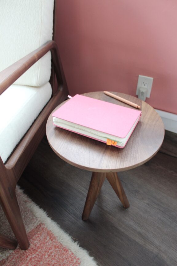 A pink notebook and a pen rest on a round wooden side table next to a chair with a white cushion.