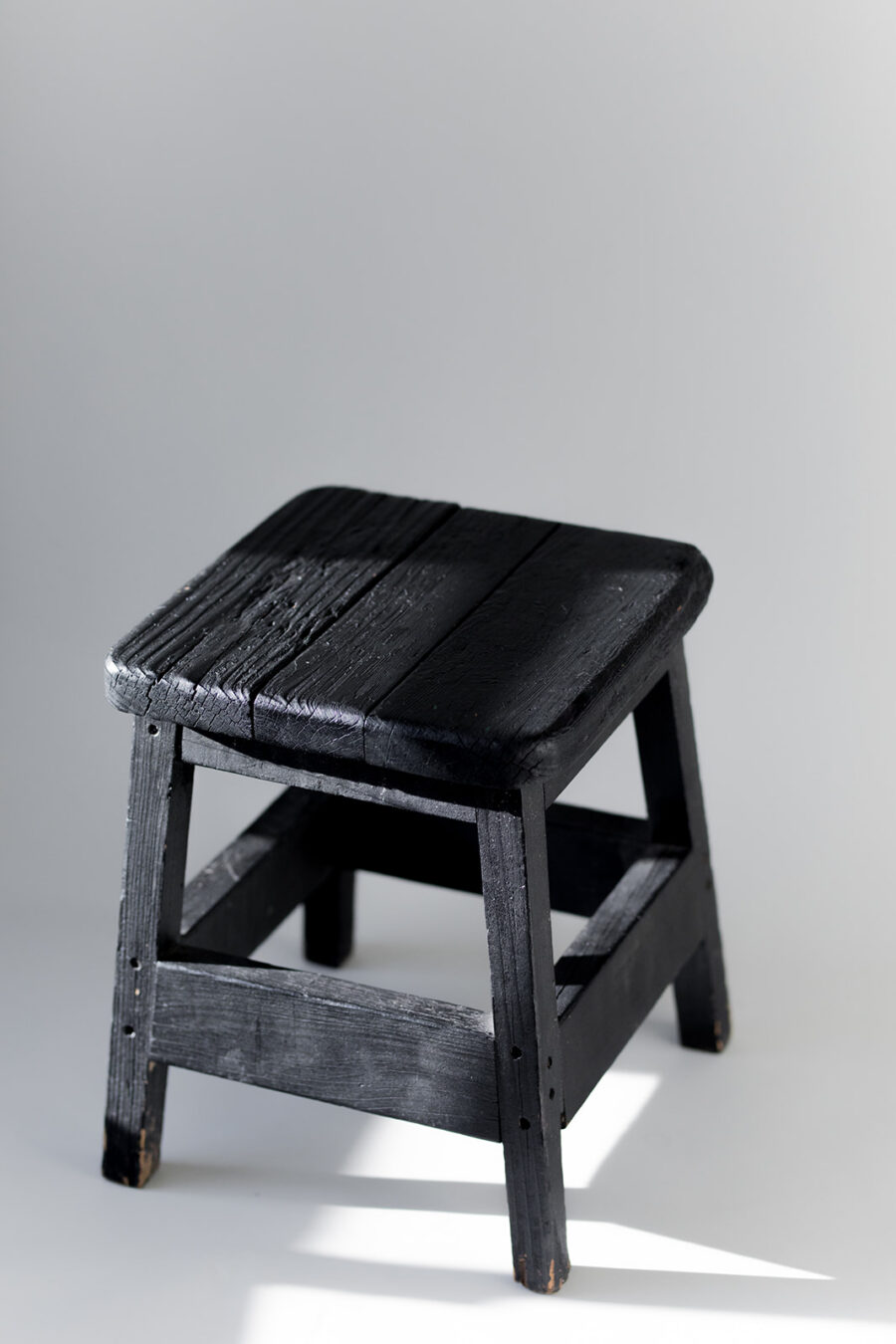 A small black wooden stool with a square seat and visible grain texture, placed on a light background with soft shadows.