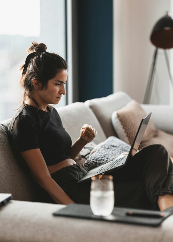 Woman sitting on a couch with a laptop on her lap, focusing on the screen. A glass of water is on the table in the foreground.