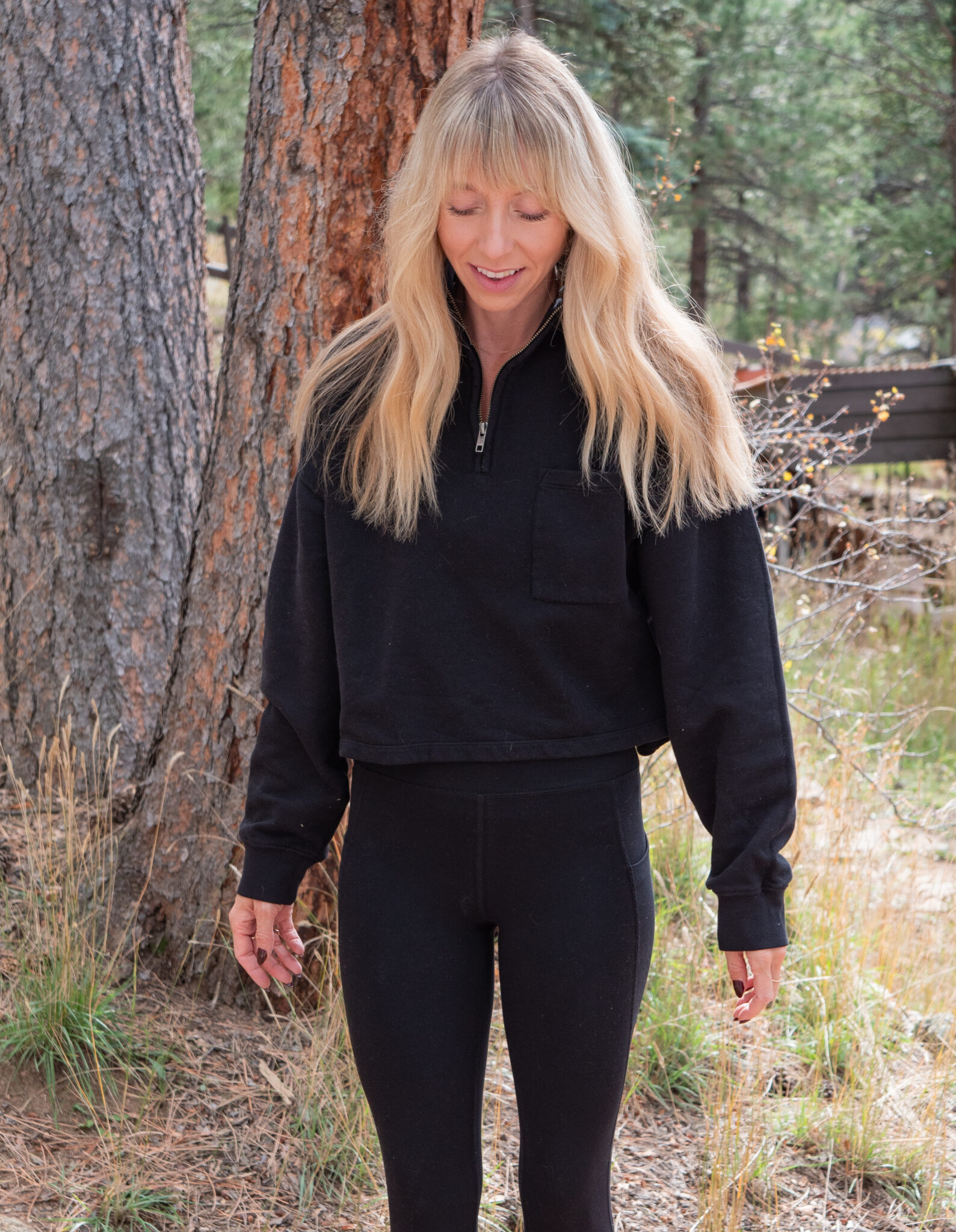 Woman with long blonde hair wearing a black zip-up sweater and black leggings stands outdoors near trees and grass, looking down with a slight smile.