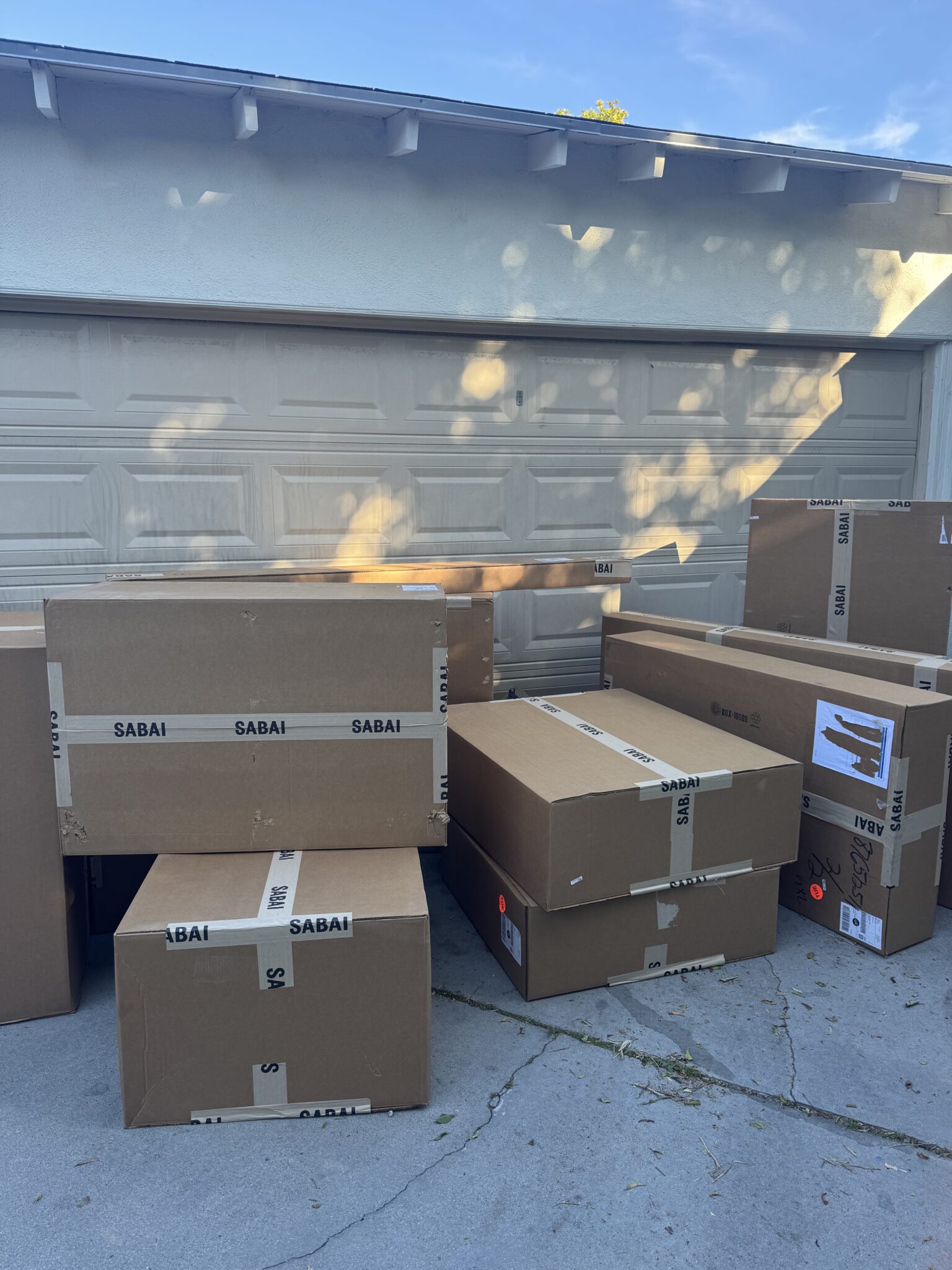 Several large cardboard boxes labeled "SABAI" are stacked in front of a closed garage door on a driveway.