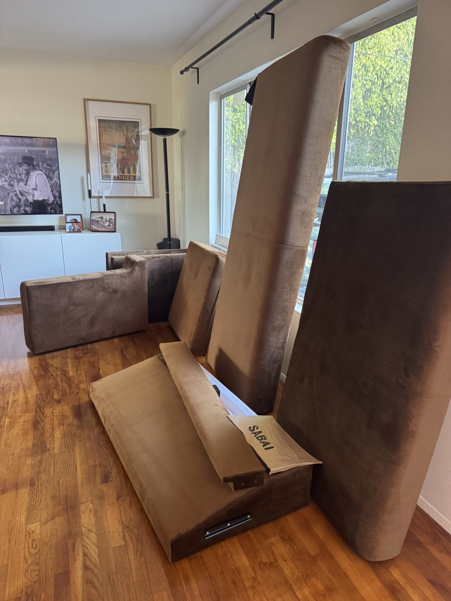 Several large brown couch cushions and parts are propped against a living room wall on hardwood floors next to a window.