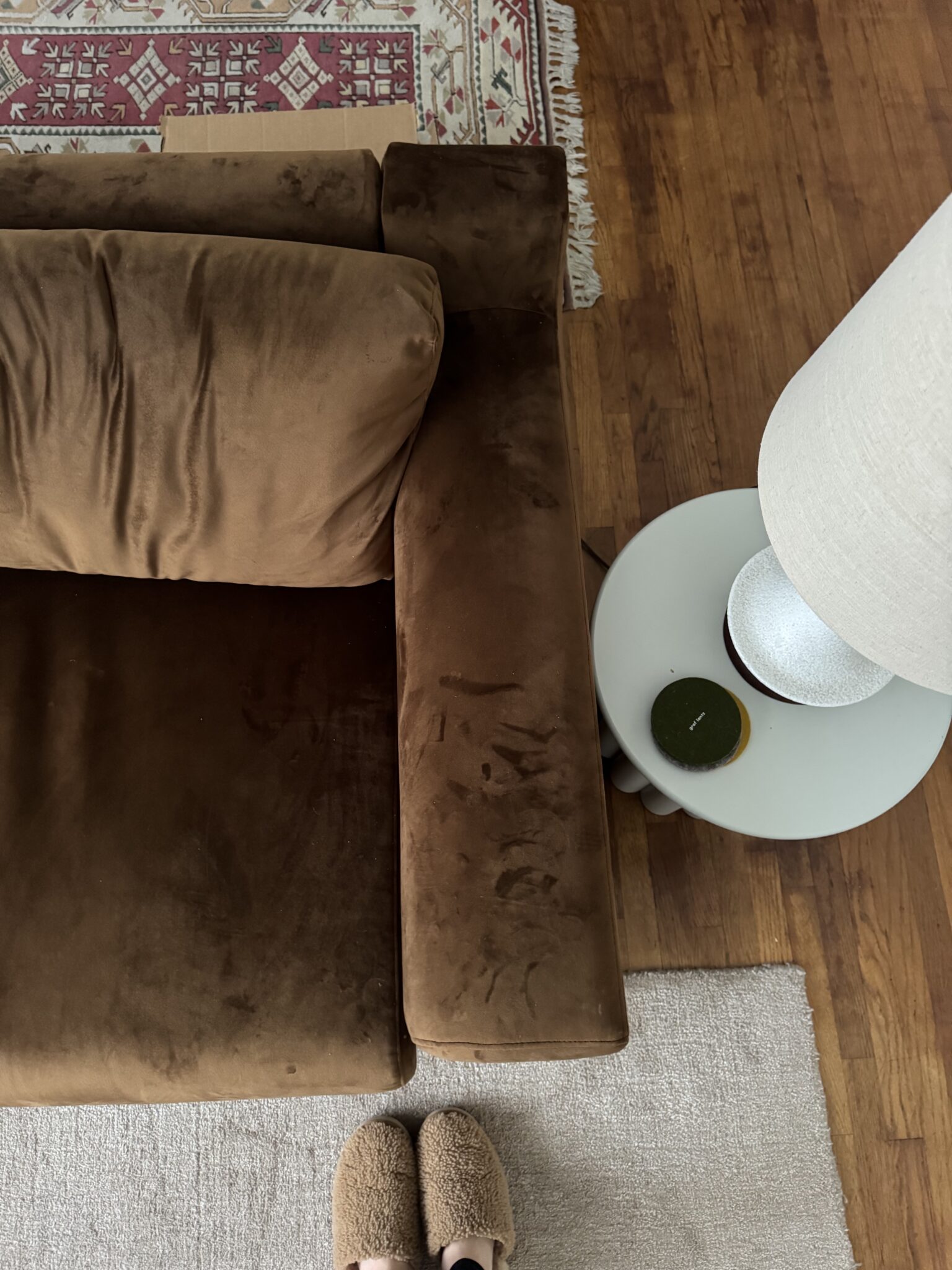 Brown velvet sofa next to a white round side table with a lamp and green coaster on it; part of a rug and slippers visible on a wooden floor.
