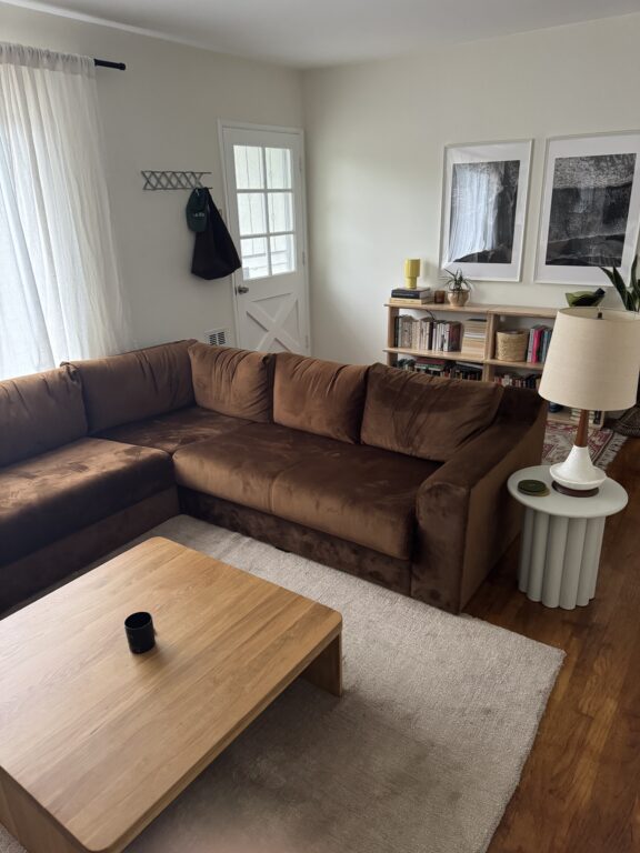 A living room with a brown sectional sofa, wooden coffee table, white side table with a lamp, bookshelf, framed art, and a door leading outside.