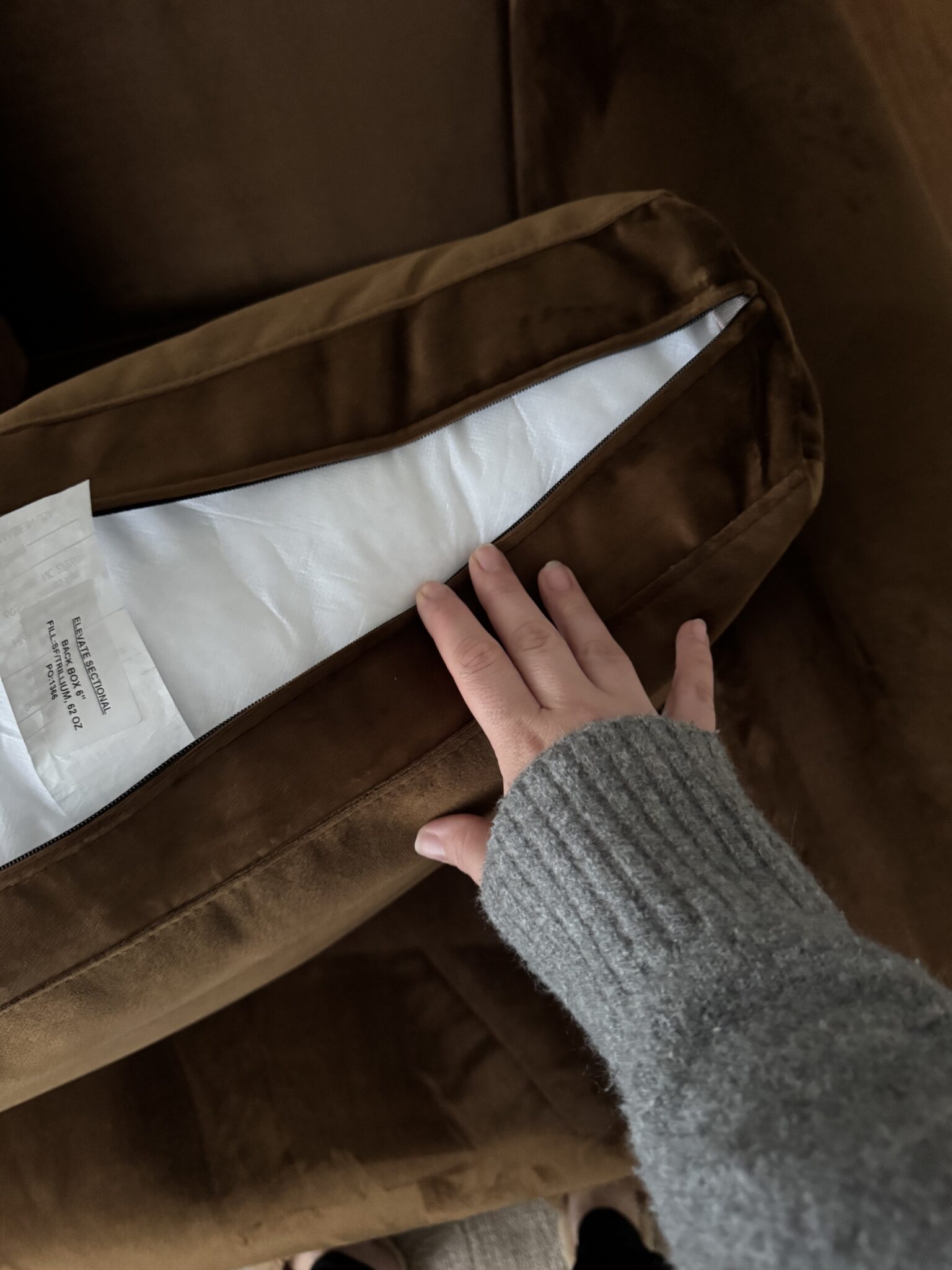 A hand in a gray sweater opens the zipper of a brown cushion to reveal the white inner pillow and a visible care label.