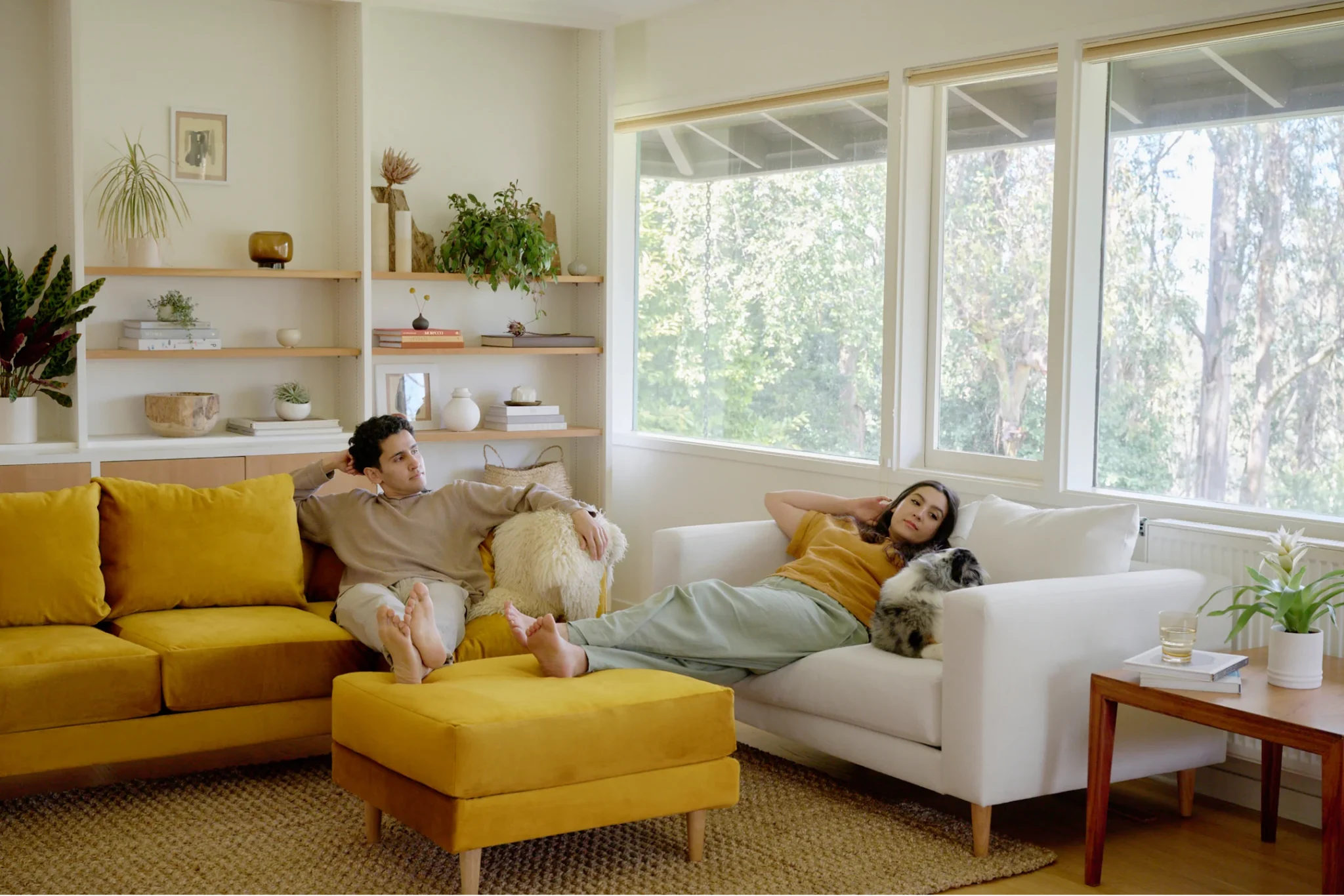 Two people relax in a bright living room, one sitting on a yellow sofa and the other lying on a white sofa with a small dog. Large windows show trees outside.