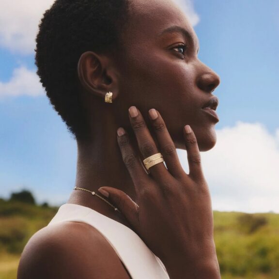 A person with short hair poses outdoors, wearing gold earrings, a ring, and a necklace, with one hand touching their face against a blue sky background.