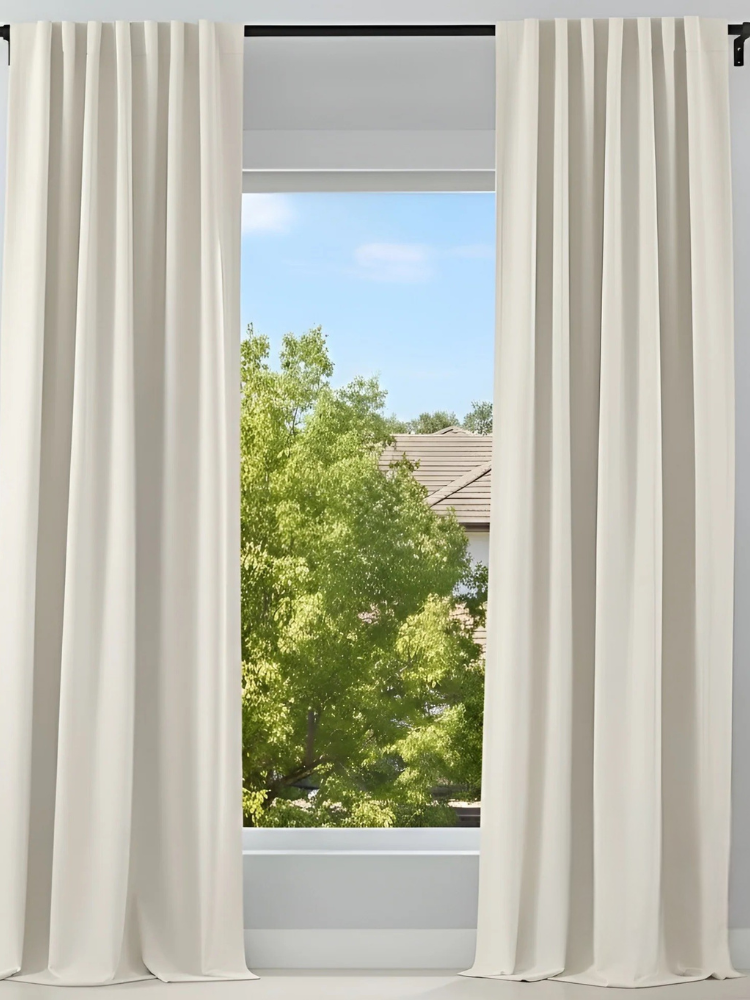 A window with beige curtains partially open, revealing a view of green trees and a house with a tiled roof outside.