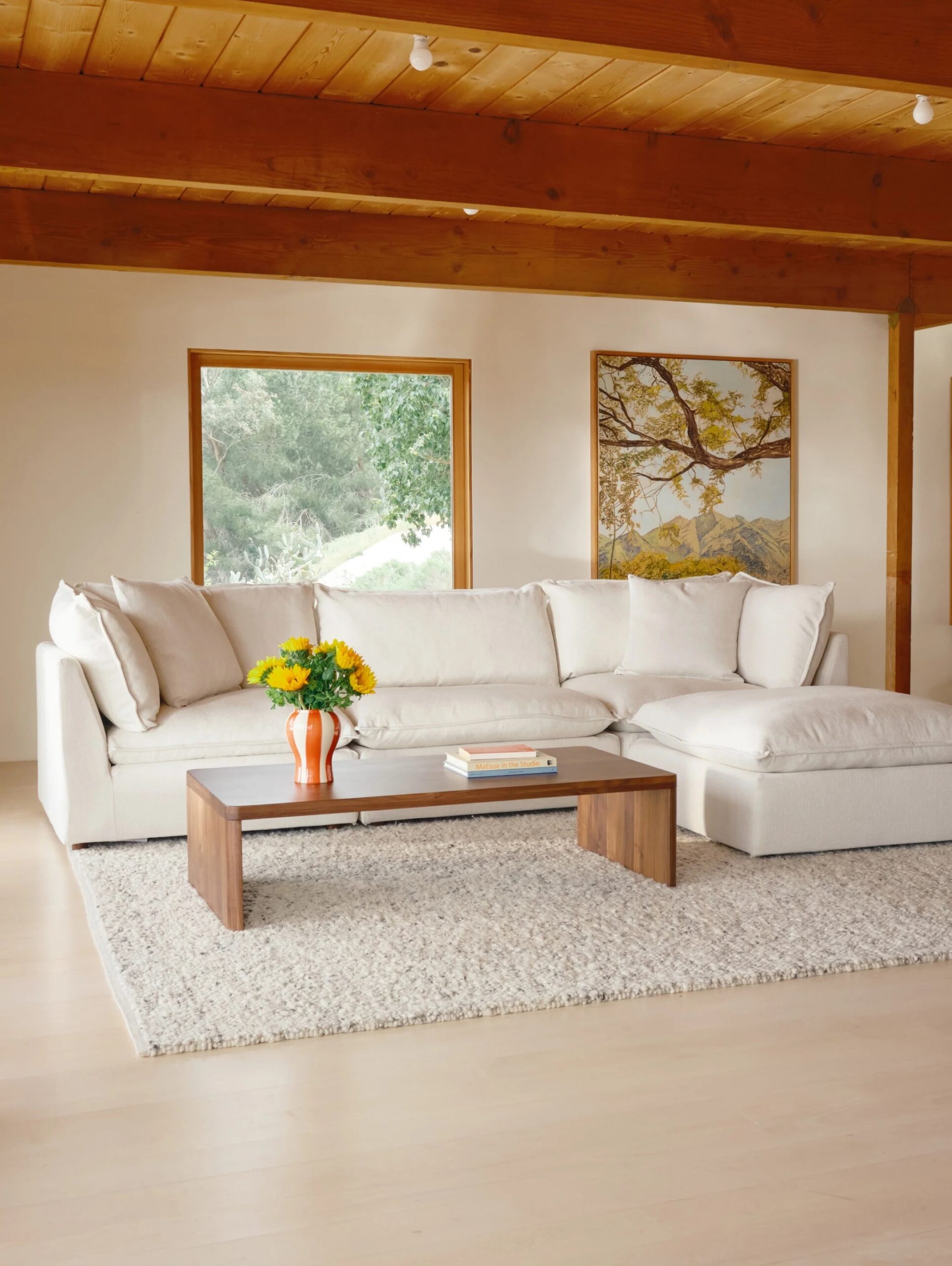 A modern living room with a white sectional sofa, wooden coffee table, a vase of yellow flowers, and large windows showcasing greenery outside.
