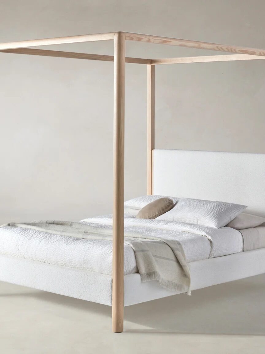 A modern canopy bed with a light wood frame and white upholstered headboard and base, dressed with white bedding, a beige throw, and a single accent pillow, set against a plain background.