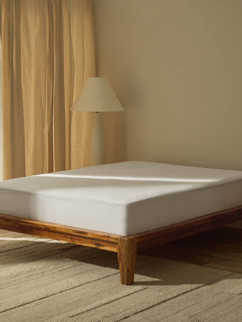 A minimalist bedroom with a wooden bed frame, plain mattress, floor lamp, beige curtains, and a neutral rug on a wooden floor.