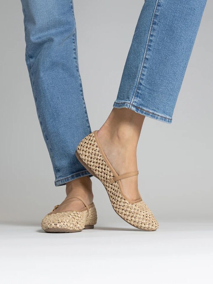 Person wearing light blue jeans and beige woven Mary Jane-style flats stands against a plain background.