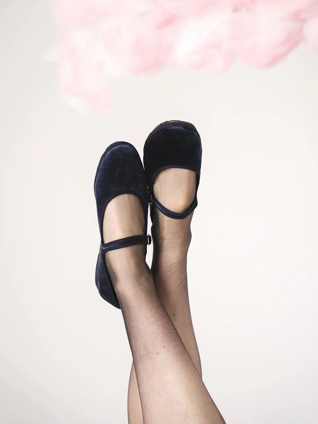 A person wearing sheer black tights and black Mary Jane shoes with their legs crossed, with pink clouds in the background.