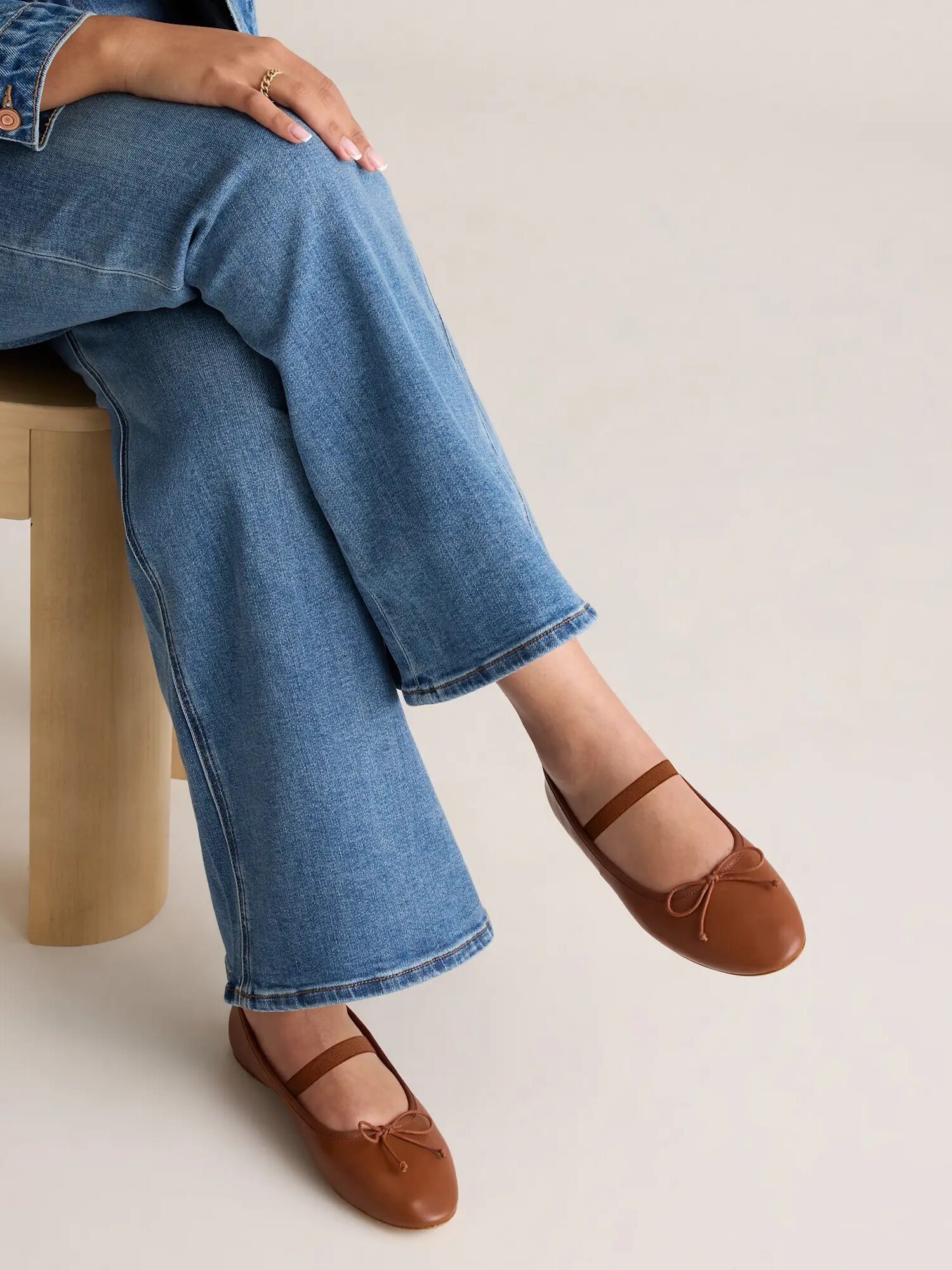 A person wearing blue jeans and brown ballet flats with a strap sits on a light wooden chair against a neutral background.