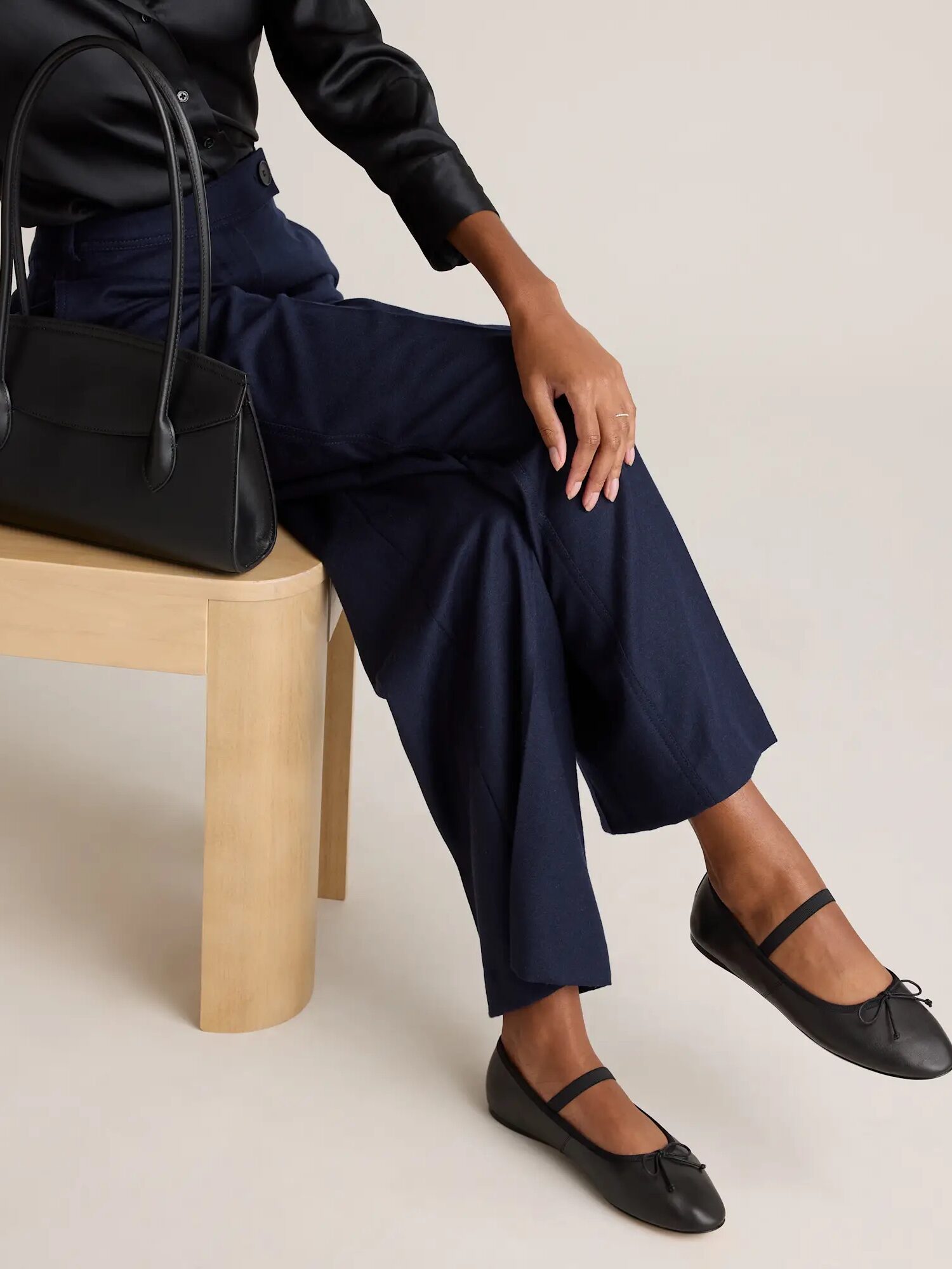 A person sitting on a wooden bench wears navy pants, a black long-sleeve top, black Mary Jane flats, and holds a black handbag.