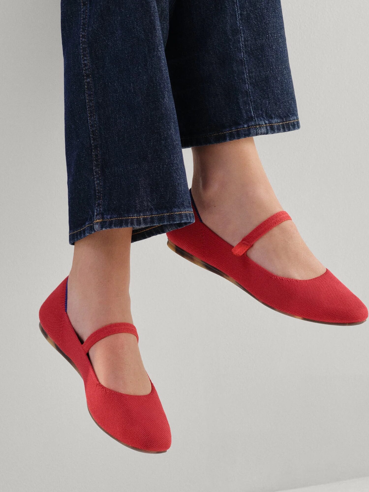 Person wearing cropped dark blue jeans and bright red Mary Jane flats, feet slightly raised against a plain light background.