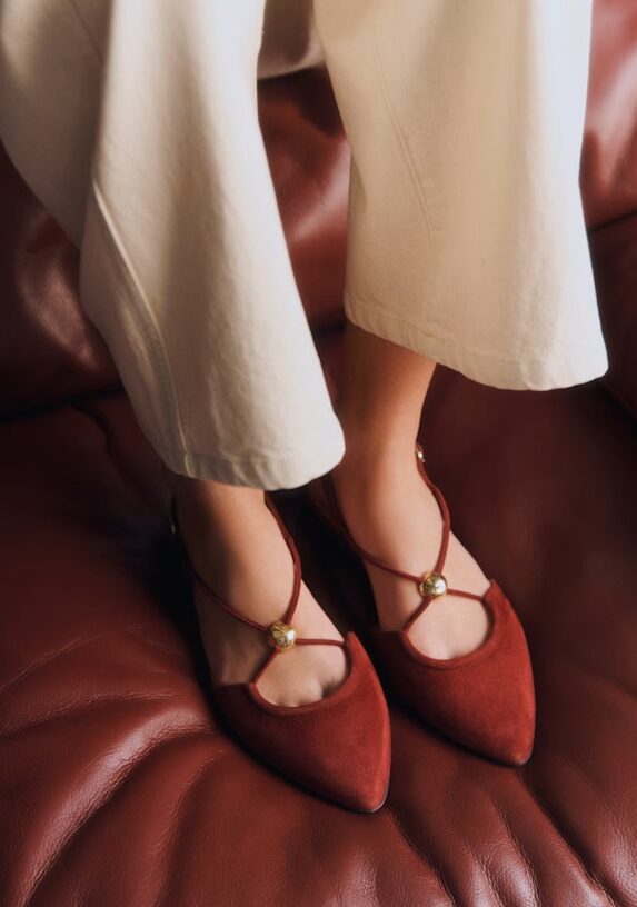 A person wearing cream-colored pants and pointed-toe, red suede flats with gold embellishments sits on a brown leather surface.