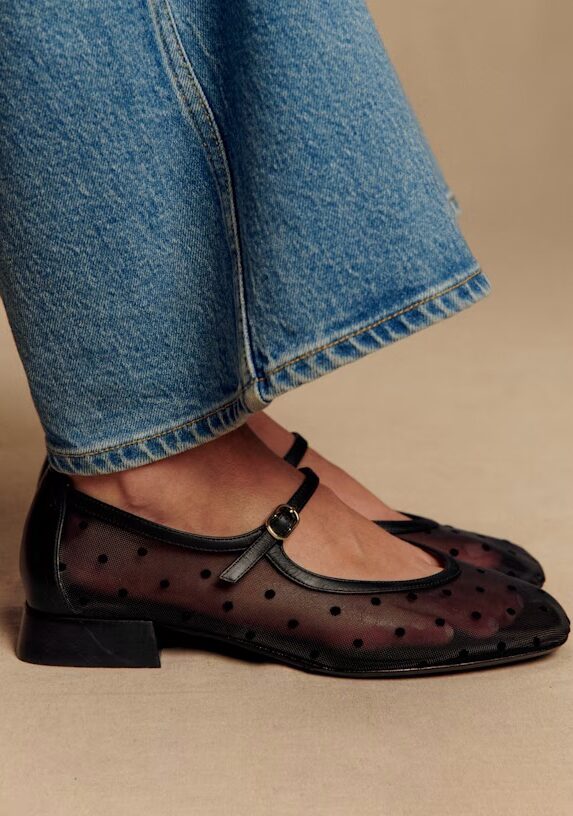 Person wearing blue jeans and black mesh Mary Jane shoes with a polka dot pattern and a low heel.