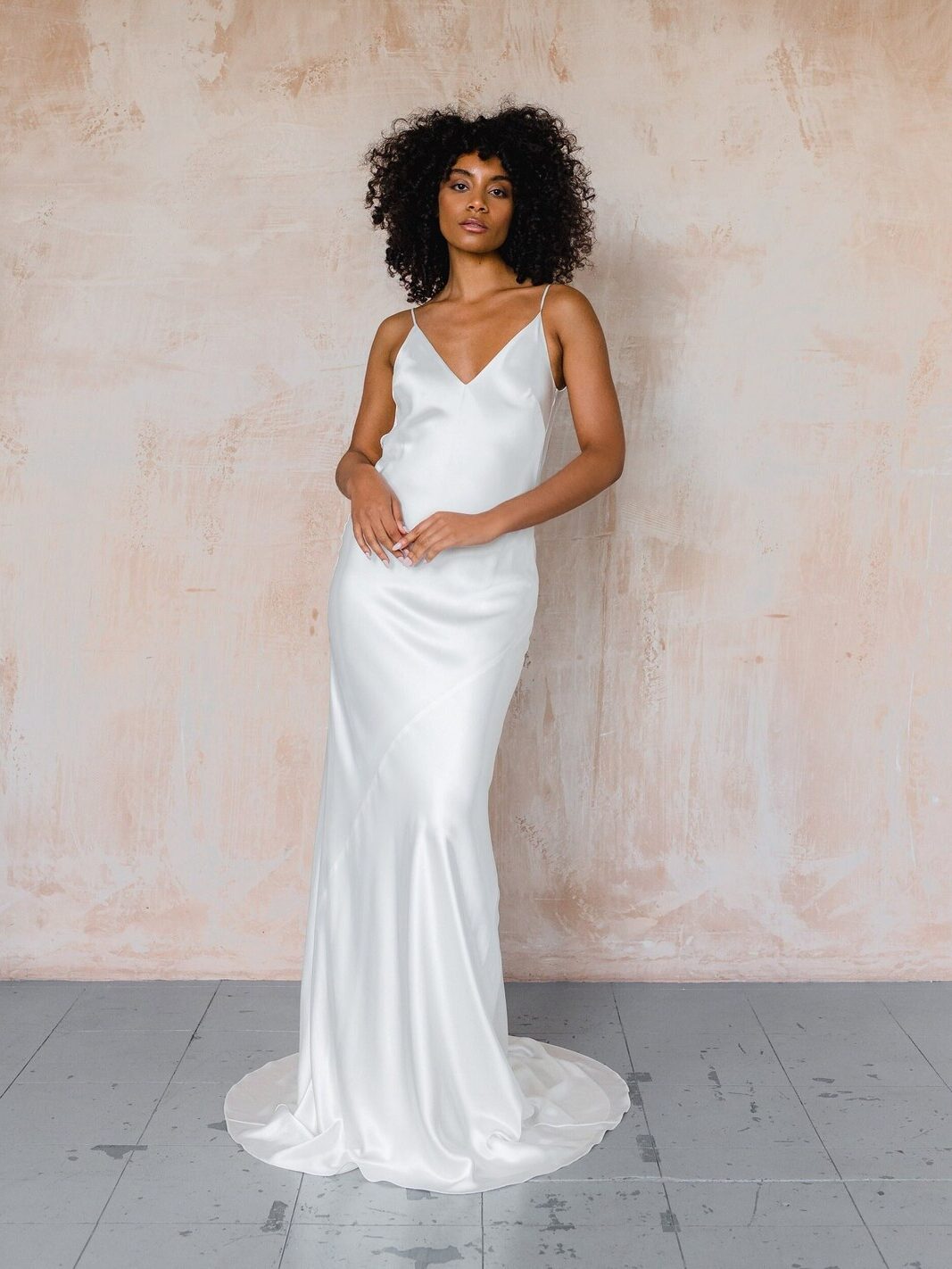 A woman wearing a white, sleeveless satin gown stands against a textured, light-colored wall on a gray tiled floor.