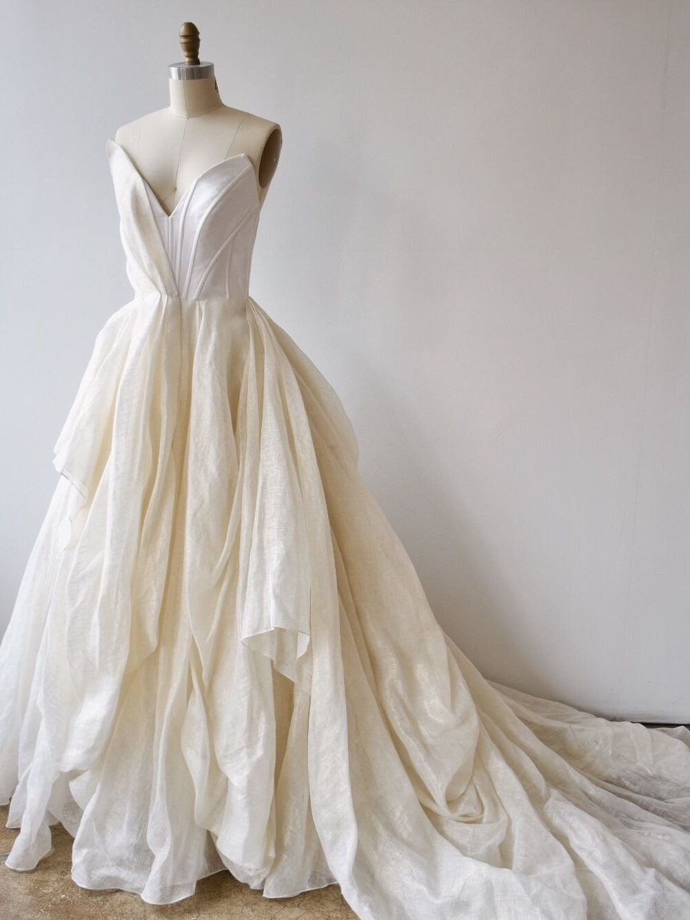 A white strapless ball gown with a deep V neckline, fitted bodice, and a full, layered skirt displayed on a dress form against a plain background.