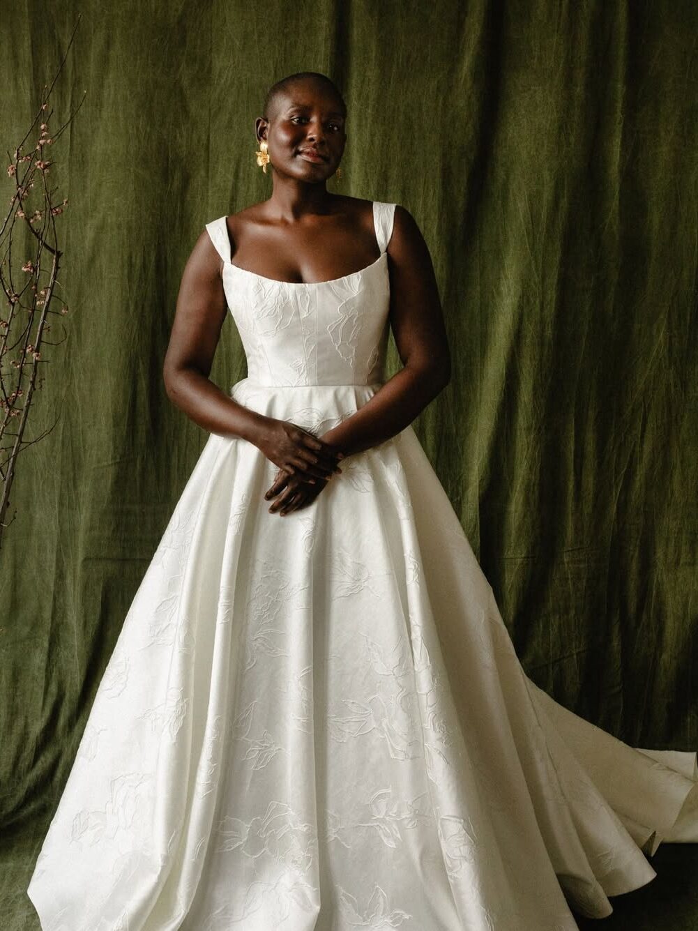 A woman stands in front of a green backdrop, wearing a sleeveless white gown with a full skirt and subtle floral patterns.