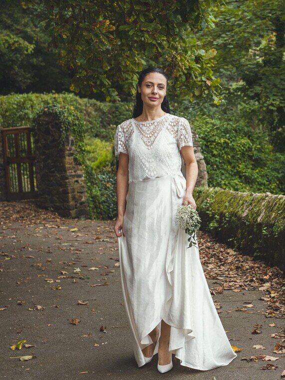 A woman in a white wedding dress holding a small bouquet stands on a leaf-strewn path beside a stone wall in a wooded outdoor setting.