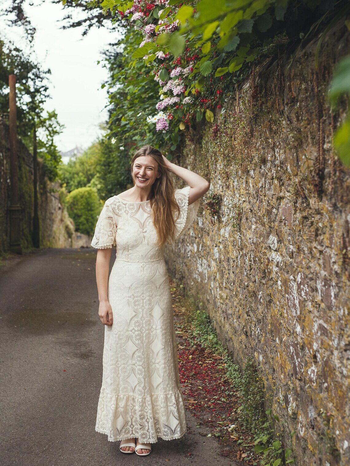 A woman in a white lace dress stands on a narrow path beside a stone wall covered with greenery and flowers, smiling with one hand in her hair.
