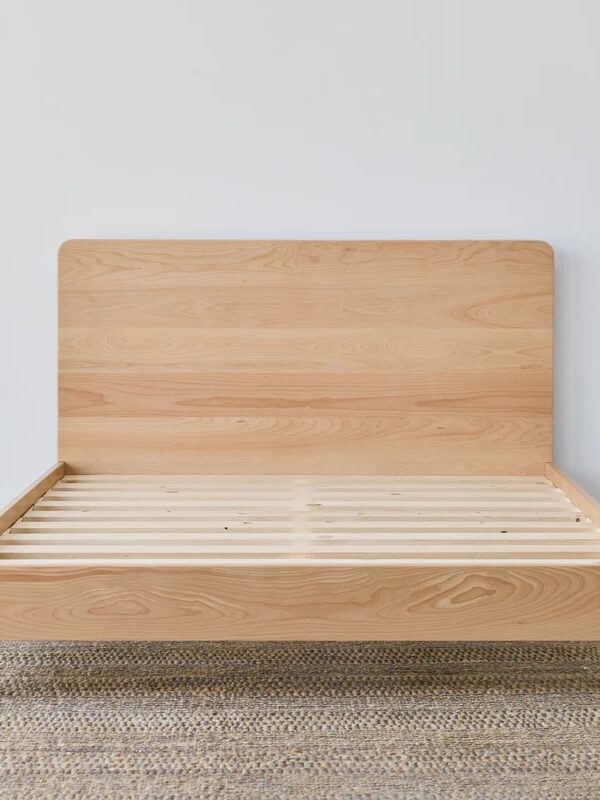 A wooden bed frame with a simple headboard and slatted base, placed on a woven rug against a plain white wall.