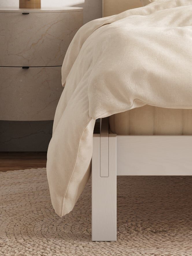 Close-up of a beige comforter on a light wood bed frame, next to a round woven rug and a marble nightstand.
