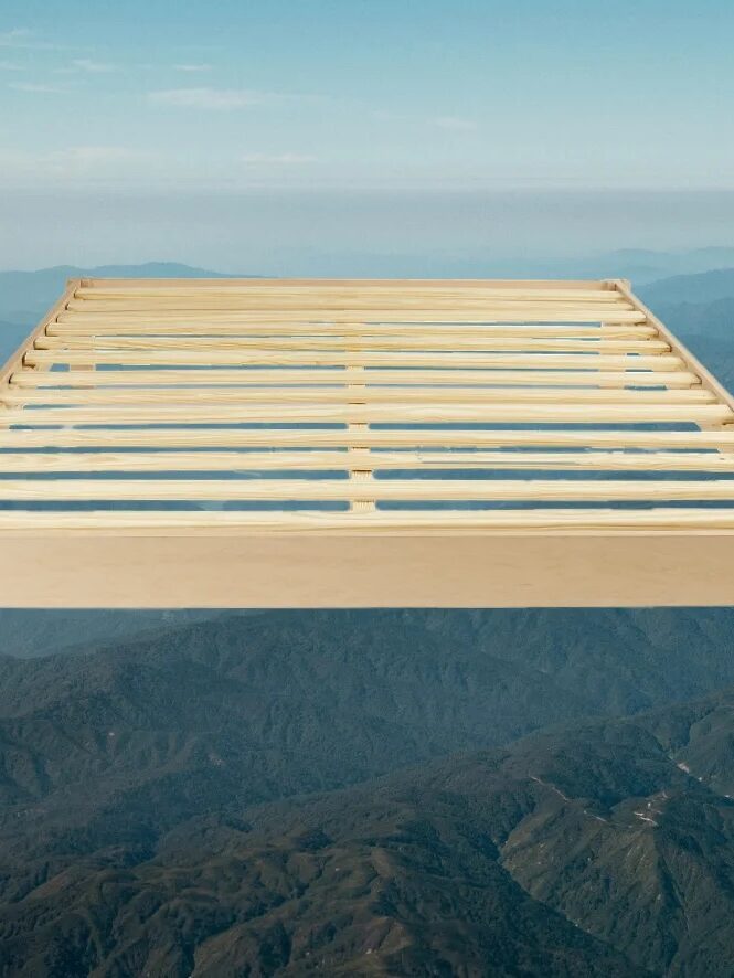 A wooden bed frame with slats is positioned in the foreground, appearing to float above a mountain landscape under a blue sky.