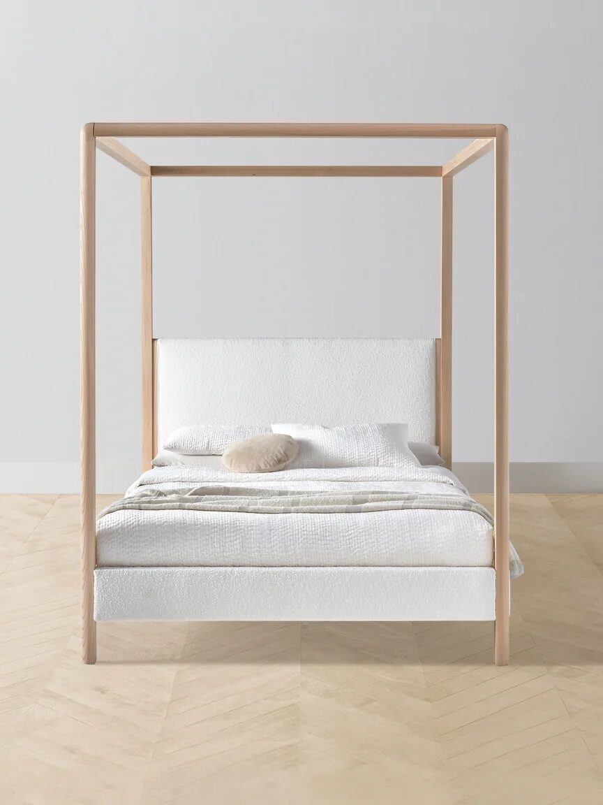 A minimalist canopy bed with a light wood frame and white bedding is positioned against a plain white wall on a light wood herringbone floor.