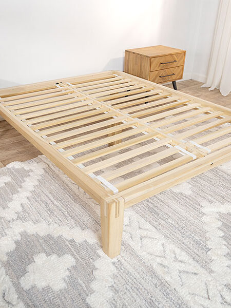 A wooden bed frame with slats is placed on a patterned rug between two wooden nightstands in a bright bedroom.