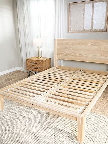 A wooden bed frame without a mattress stands between two matching wooden nightstands with lamps in a bright, minimalist bedroom.