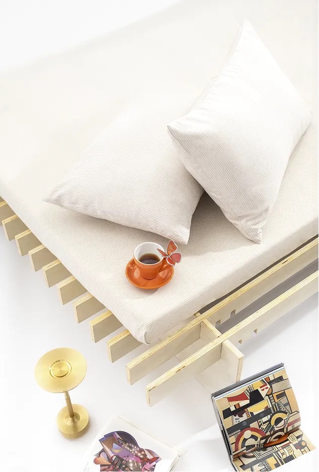 A minimal bed with a light-colored mattress and two pillows, a cup and saucer with a plant, a gold lamp, and an open book with abstract art on the floor nearby.