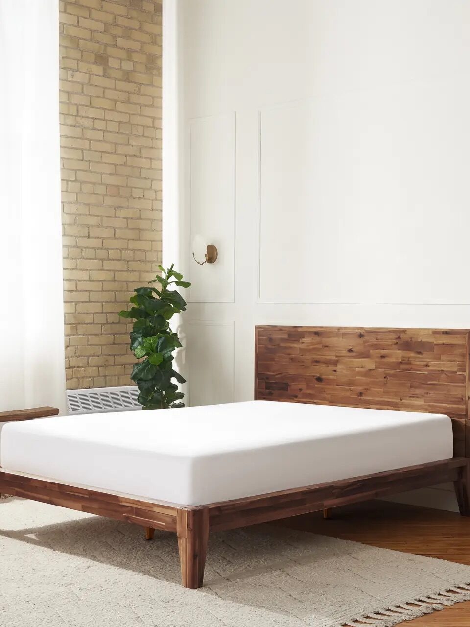 A minimalist bedroom with a wooden bed frame, a white mattress, a small wooden nightstand with a plant, and natural light coming through sheer curtains.