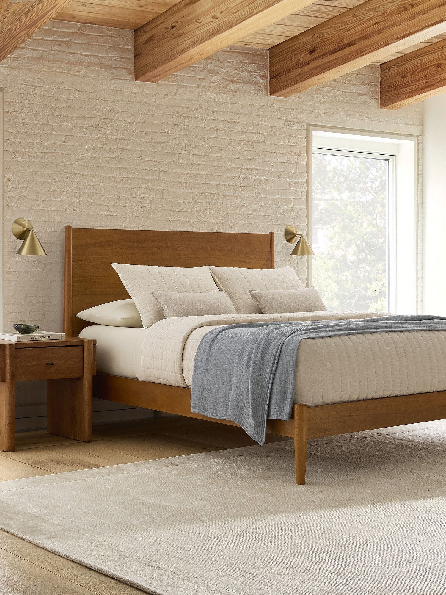 A modern bedroom with a wooden bed frame, white bedding, a gray blanket, two matching nightstands, brass wall lamps, and exposed wooden ceiling beams.
