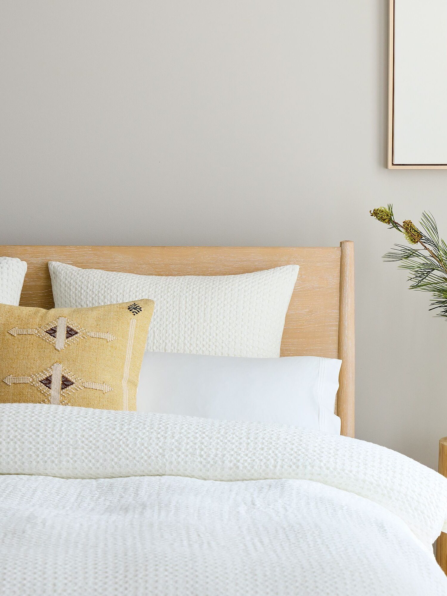 A neatly made bed with white bedding, a yellow decorative pillow, a wooden headboard, and a side table holding a vase with greenery; framed art hangs on a light wall.