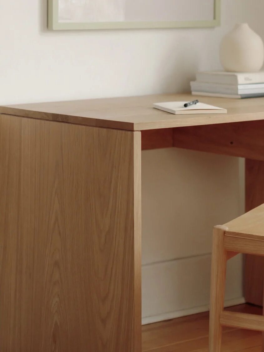 A wooden desk with a matching chair, a notepad and pen, two stacked books, and a round white vase against a white wall.