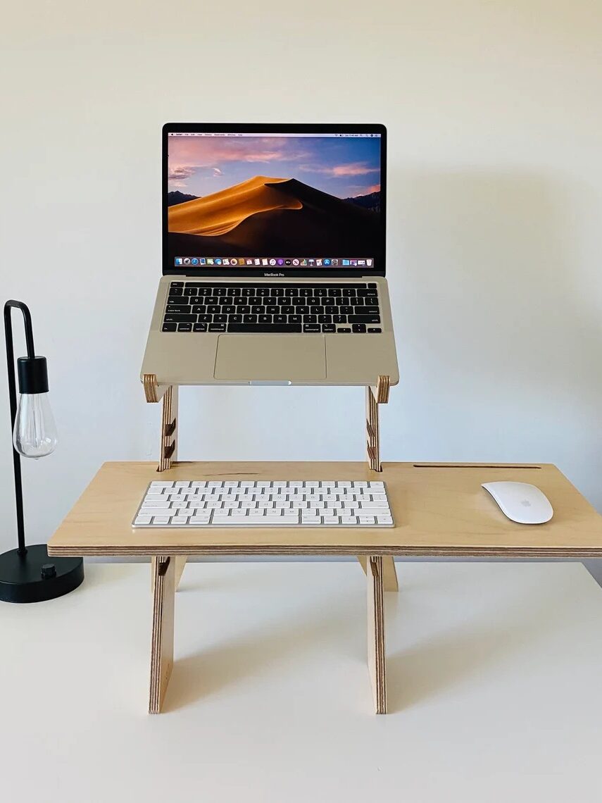 A minimalist wooden standing desk holds a laptop, keyboard, and mouse on a white table next to a black desk lamp.