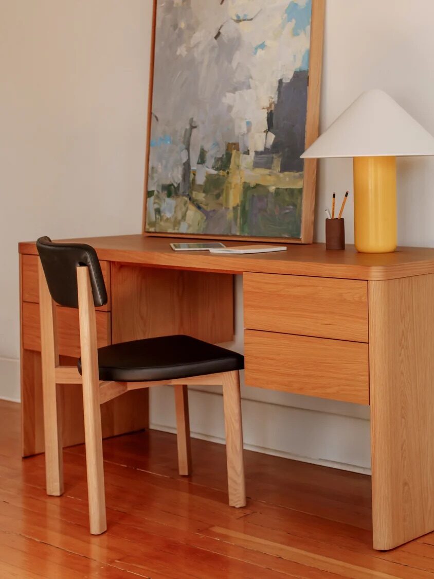 A wooden desk with a chair, lamp, painting, and writing tools on top. One drawer is shown open in a close-up on the right side of the image.