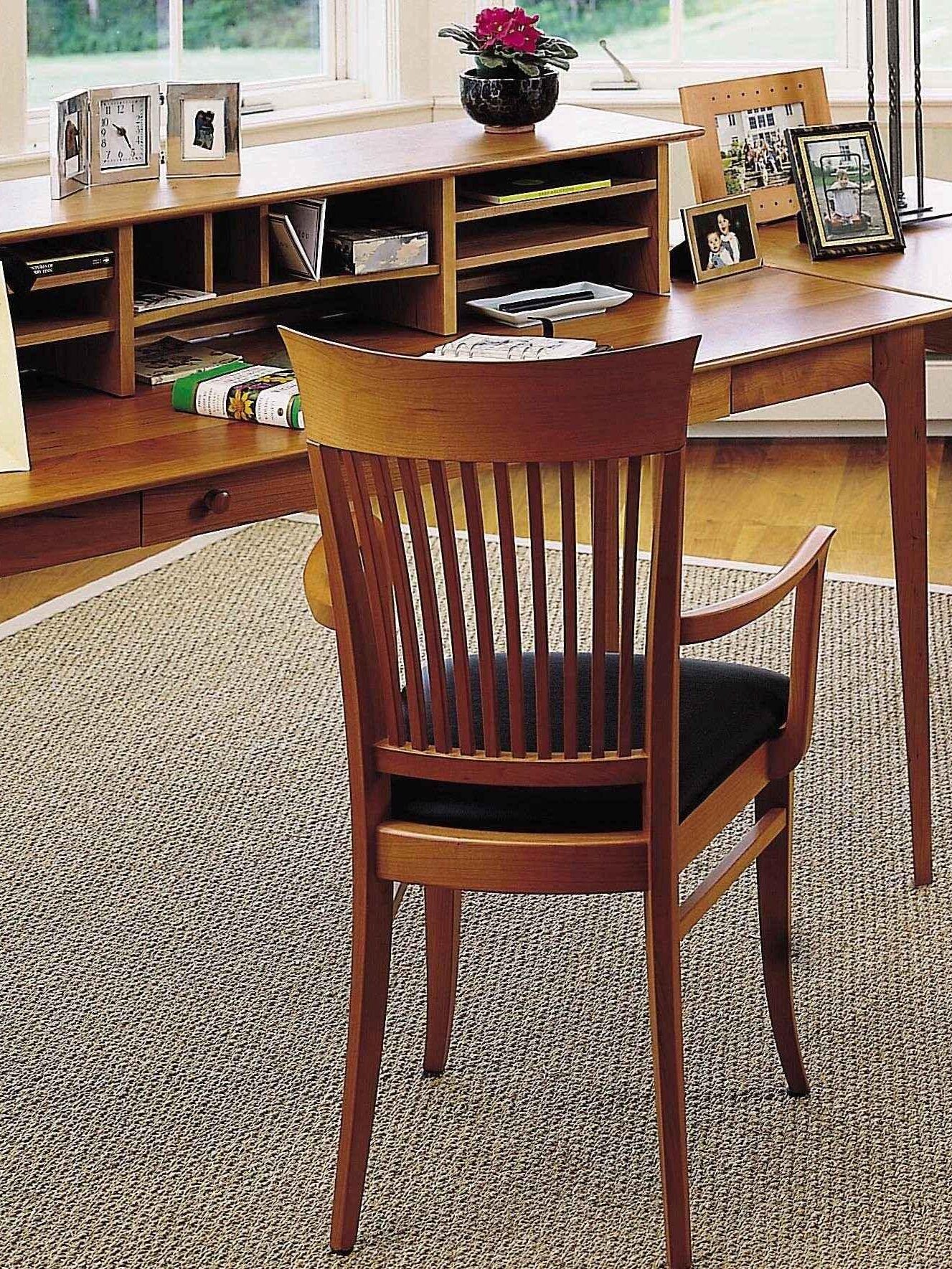 A wooden desk with shelves and a wooden chair on a woven rug; the desk holds office supplies, framed photos, a plant, and a laptop near a window.