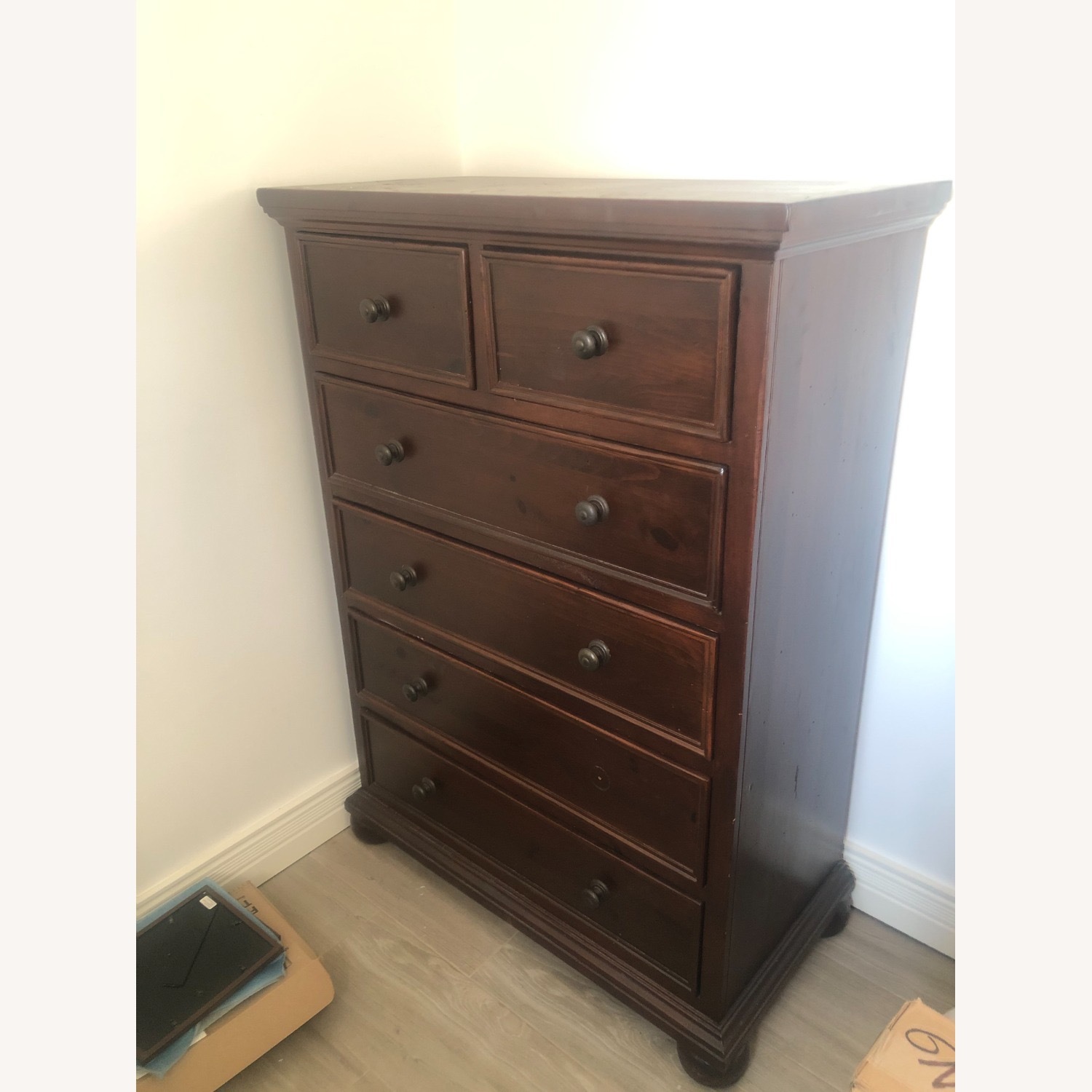 A dark brown wooden chest of drawers with six drawers, positioned against a white wall on a light-colored floor.