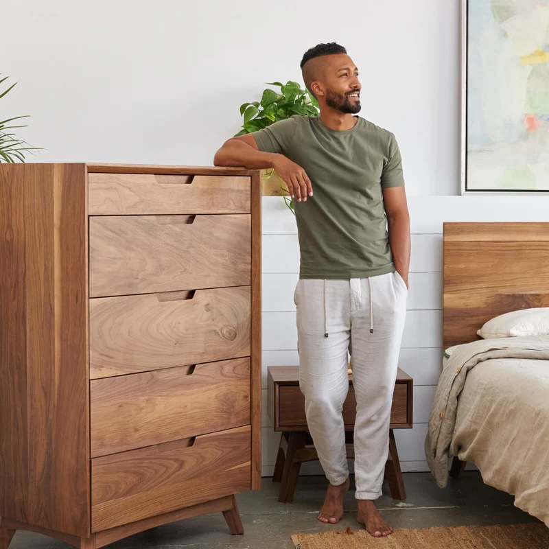 Man standing barefoot in a bedroom, leaning on a wooden dresser, wearing a green t-shirt and gray jogger pants, with a bed and plant visible in the background.