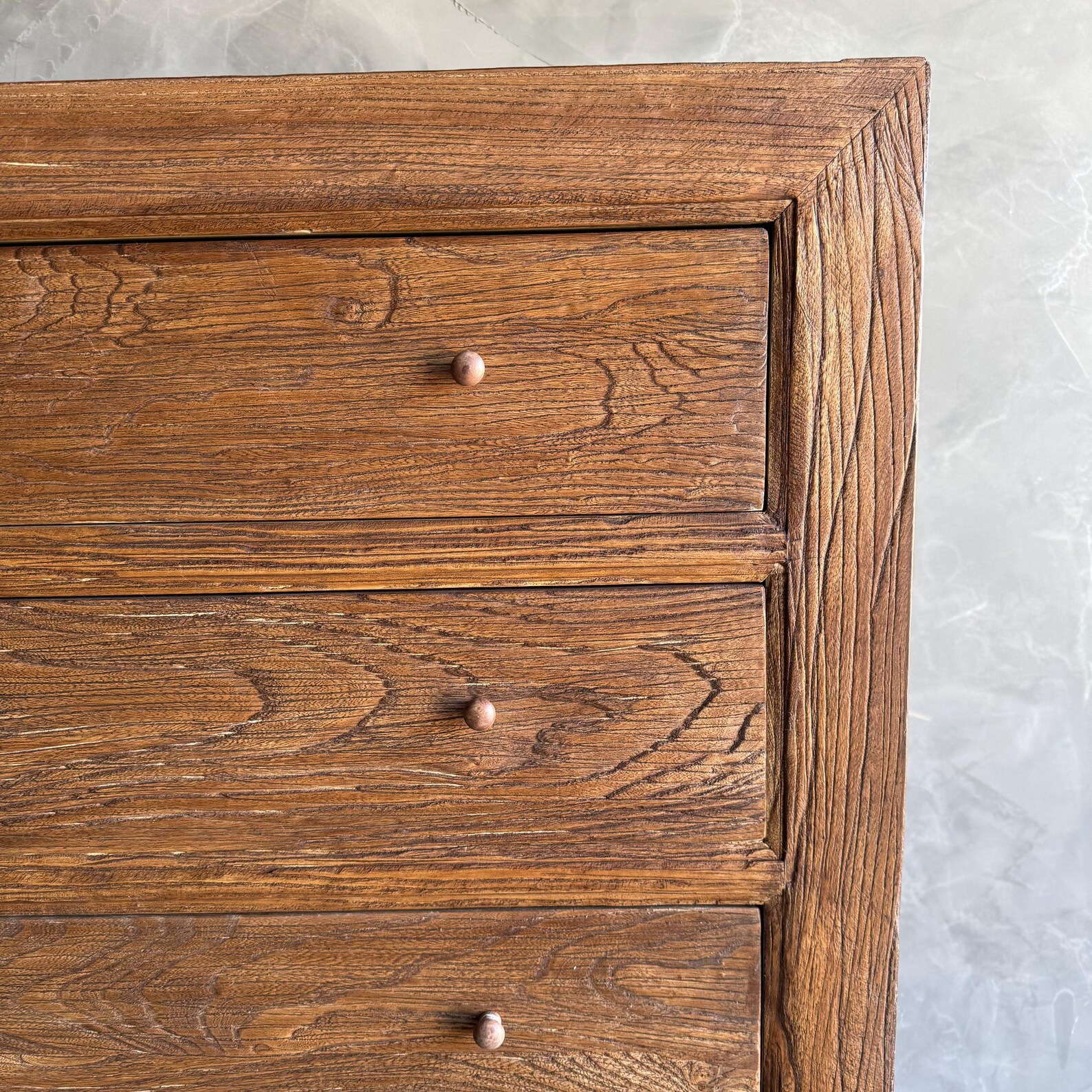Close-up of a wooden dresser with three visible drawers, each featuring a small round knob, set against a textured gray wall.
