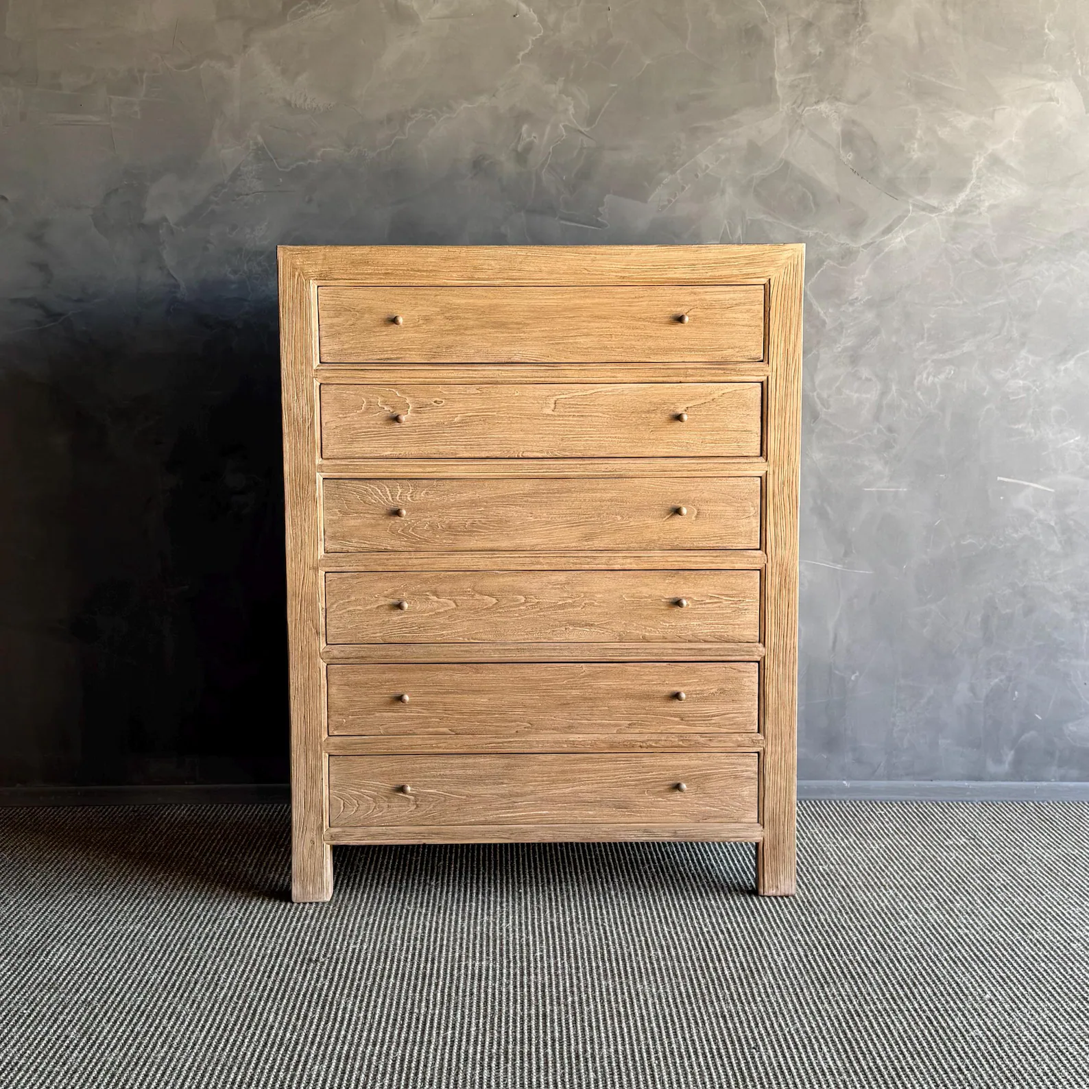 A light wood five-drawer dresser stands on a textured gray carpet in front of a smooth, dark gray wall.