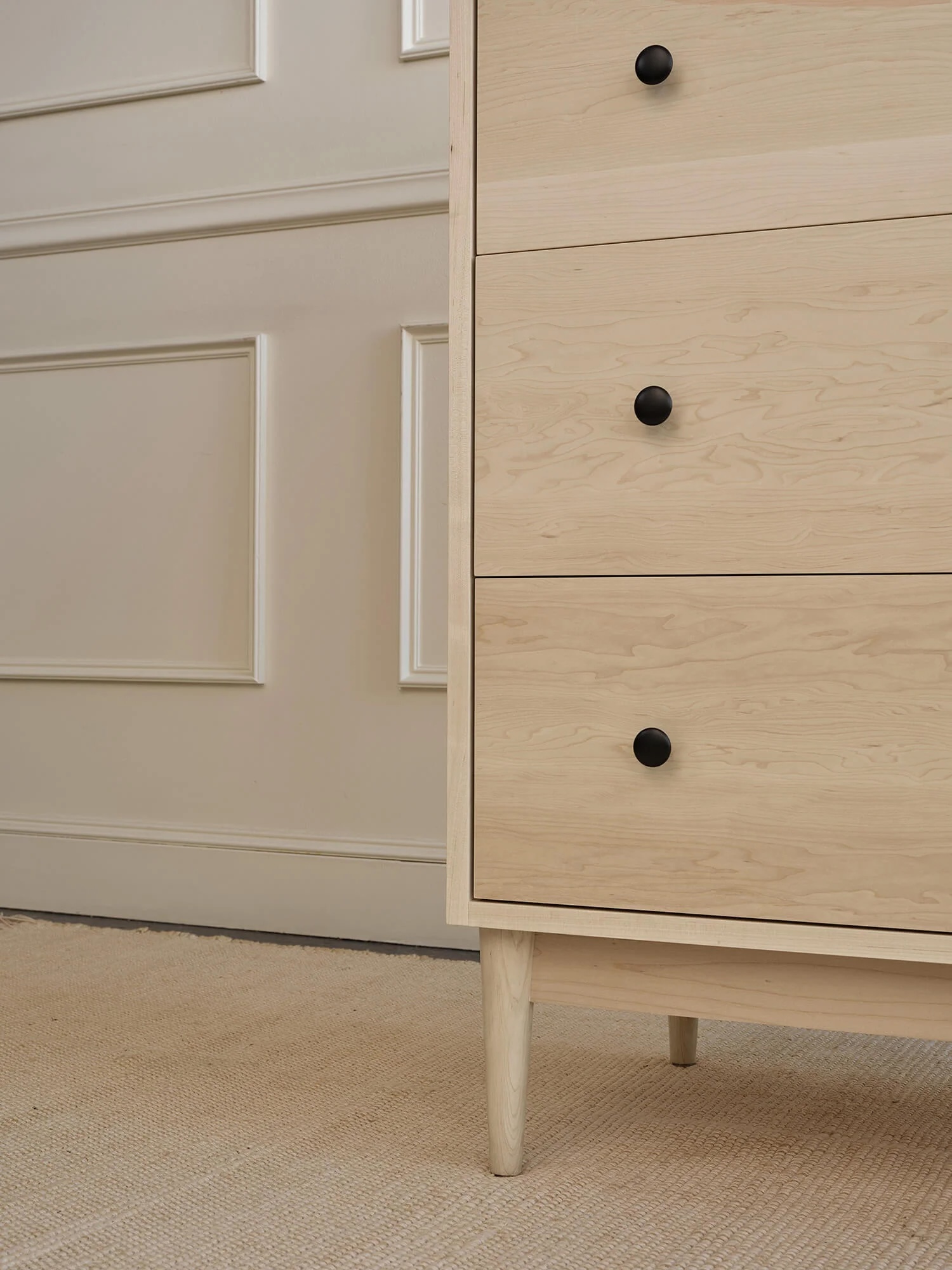 A light wood dresser with three drawers and black knobs stands on a beige carpet next to a white paneled wall.