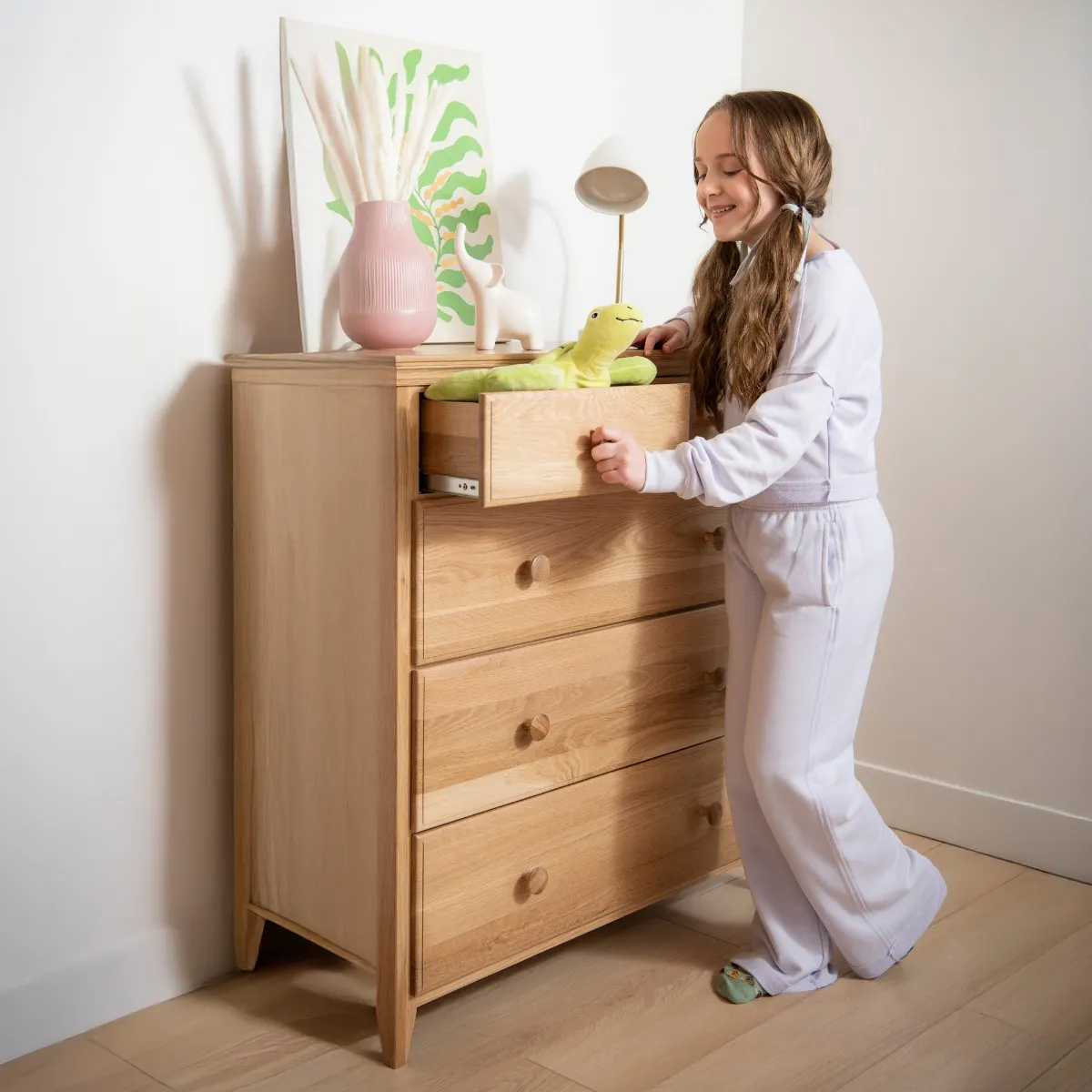 A young girl in light pajamas places a green stuffed animal inside the top drawer of a wooden dresser in a bright, modern bedroom.