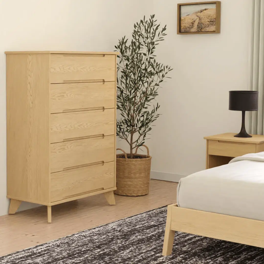 A light wood dresser with five drawers stands next to a potted plant in a bedroom with a matching nightstand, bed, and a lamp on the nightstand.